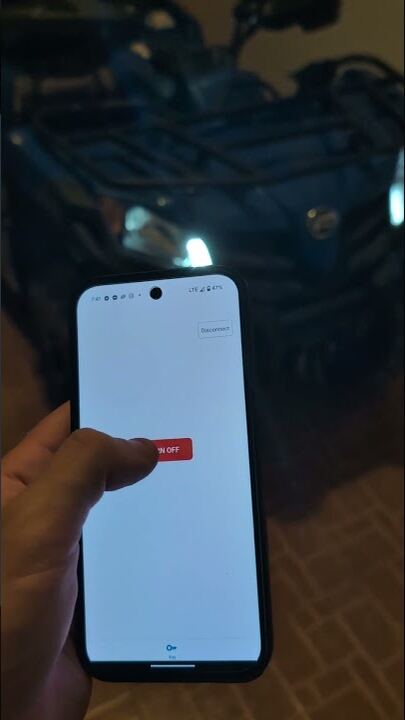 A hand holding a phone running the Keyvo app, its switch control on screen, with an ATV lit up in the dark behind it.